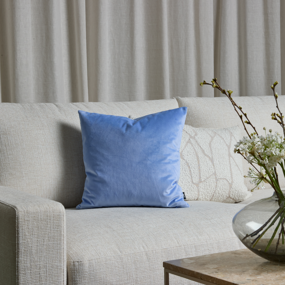 Lys beige sofa med lyseblå velourpude og glasvase med hvide blomster på stenbord foran lyse gardiner.