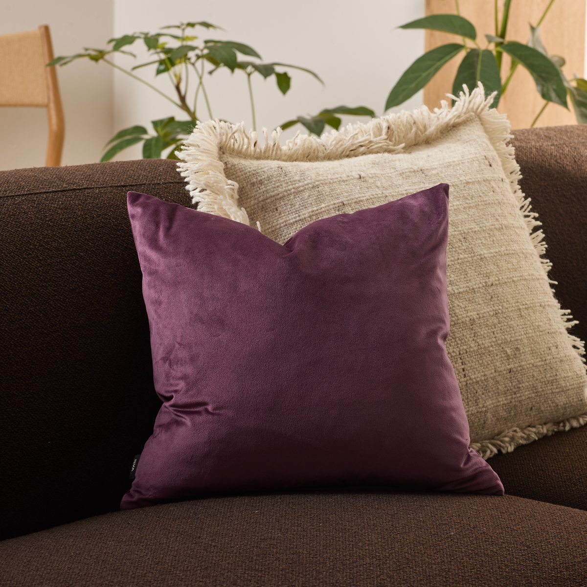 Blommefarvet pyntepude på mørkebrun sofa foran beige pude med frynser; planter i baggrunden.