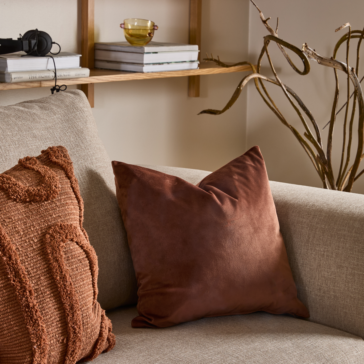 Beige sofa med to rustbrune pyntepuder; træhylde med bøger, hovedtelefoner og en lille grøn glasskål i baggrunden samt dekorative tørrede grene.