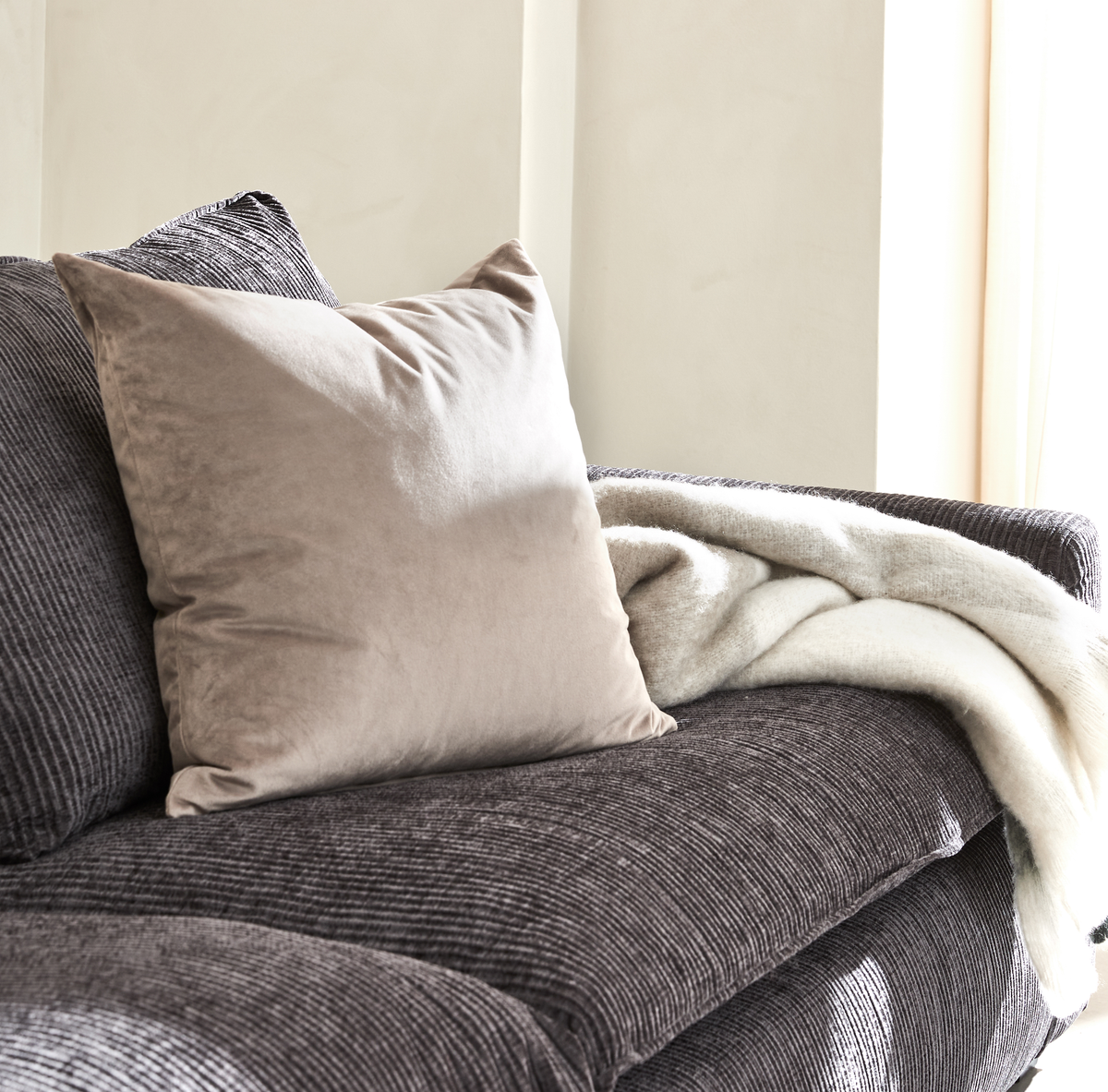 Beige velourpude på mørk rillet sofa med blød hvid plaid i dagslys ved vindue.