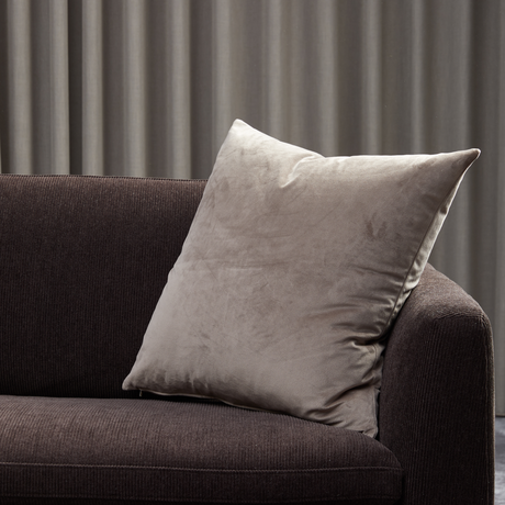 Beige velourpude lænet mod mørkebrun sofa foran grå gardiner.
