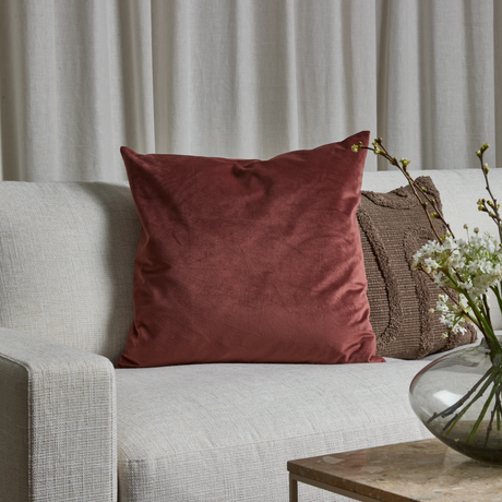 Lys sofa med stor rustrød velourpude og brun teksturpude; glasvase med hvide blomster på bord foran lyse gardiner.