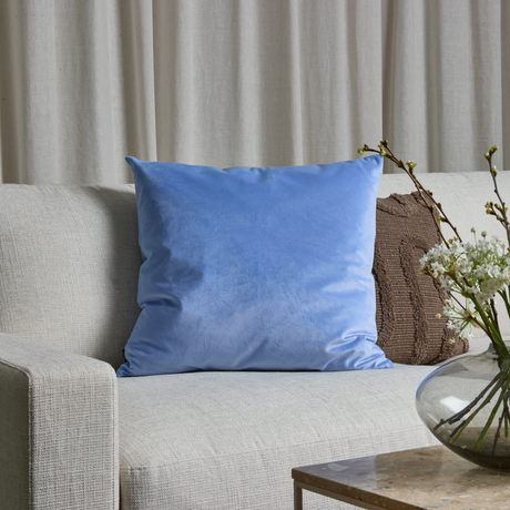 Lyseblå pyntepude på lys sofa, brun pude bagved og glasvase med hvide blomster på et sidebord.