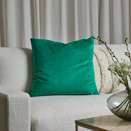 Grøn pyntepude på lys sofa med neutrale puder, beige gardiner bagved og en glasvase med hvide blomster på sofabordet.