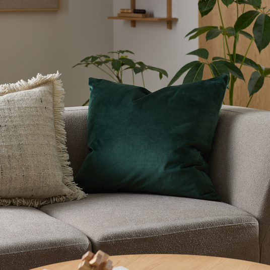Grøn velourpude på grå sofa i stue med beige frynsepude og grønne planter.