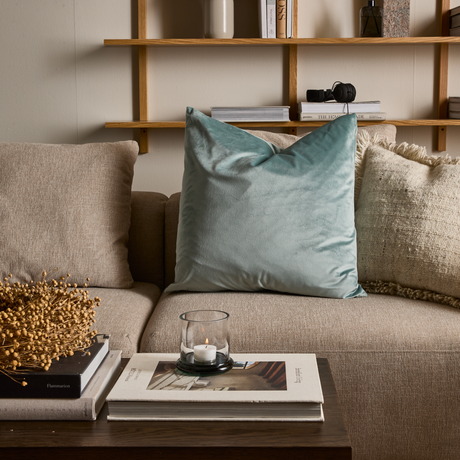 Beige sofa med mintgrøn velourpude; sofabord med glasstage på stakkede bøger, bl.a. Flammarion; væghylde i træ med vase og bøger, bl.a. The House of Jade.