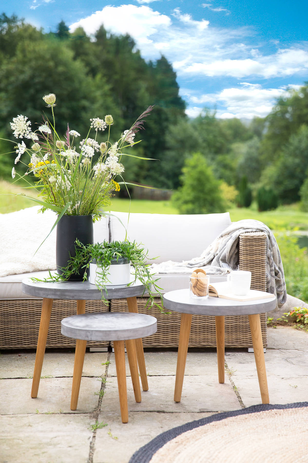 Sommerlig terrasse med rottingsofa, tre små runde borde med betontop og træben, vilde blomster i sort vase, grøn potteplante, blød plaid, bog, kop og glas med småkager foran grønne træer og blå himmel.