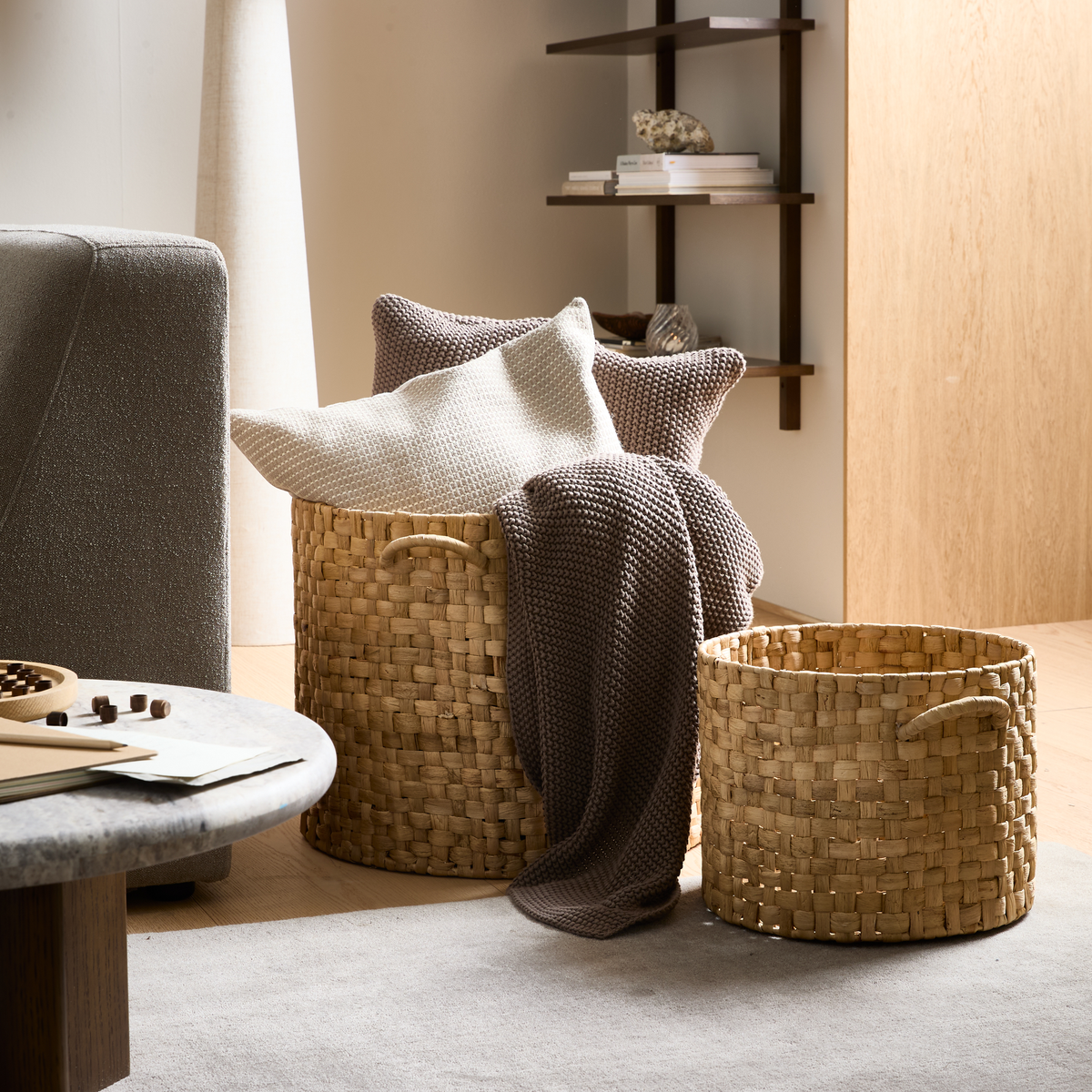 To flettede opbevaringskurve i naturmateriale med puder og en strikket plaid ved siden af en sofa og sofabord i en lys stue.