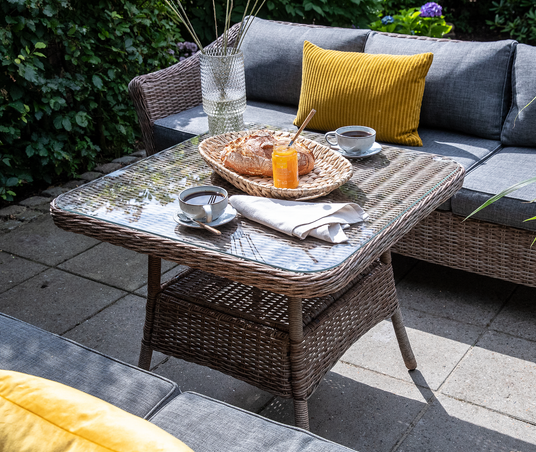 Flettet havebord med glas, grå loungesofa og gul pude; morgenmad med kaffe, brød og marmelade på terrasse i dagslys.