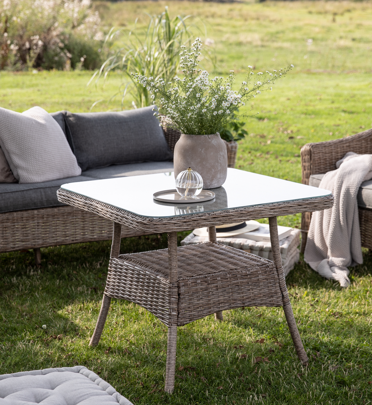 Firkantet havebord i polyrattan med glasplade, vase med hvide blomster og lille bakke, omgivet af loungesofa med puder i grøn have.