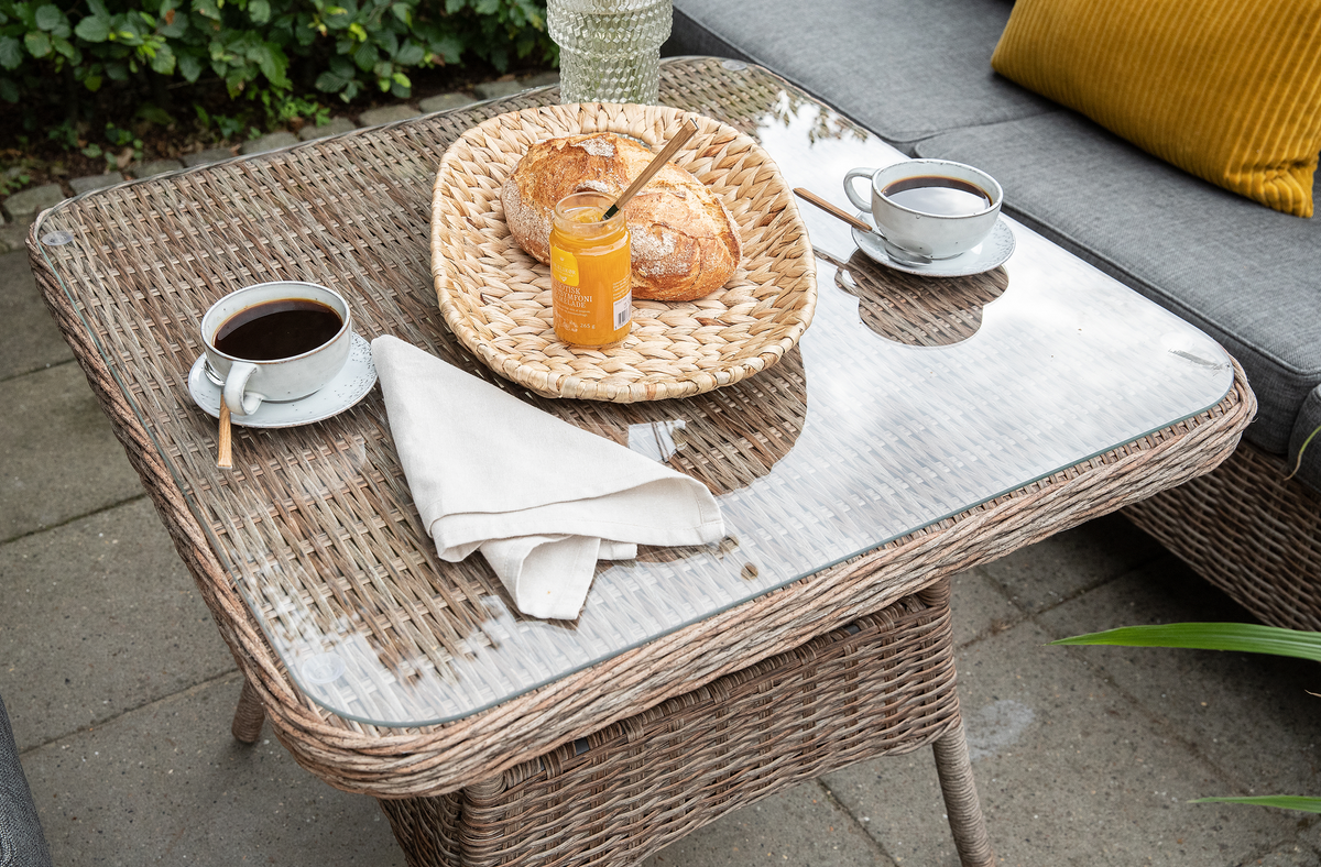 Udendørs fletbord med glasplade med to kopper kaffe, en flettet bakke med brød og et glas marmelade samt en hvid serviet; grå sofa med gul pude i baggrunden.