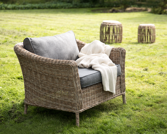 Flettet lænestol i rattan med grå hynder og cremefarvet strikket plaid på en græsplæne; to runde rattan skamler i baggrunden.