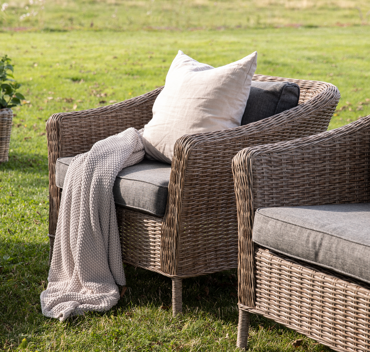 Udendørs lænestol i flet med grå hynde, beige pude og strikket plaid på græsplæne; matchende sofa ved siden af og plantekurv i baggrunden.