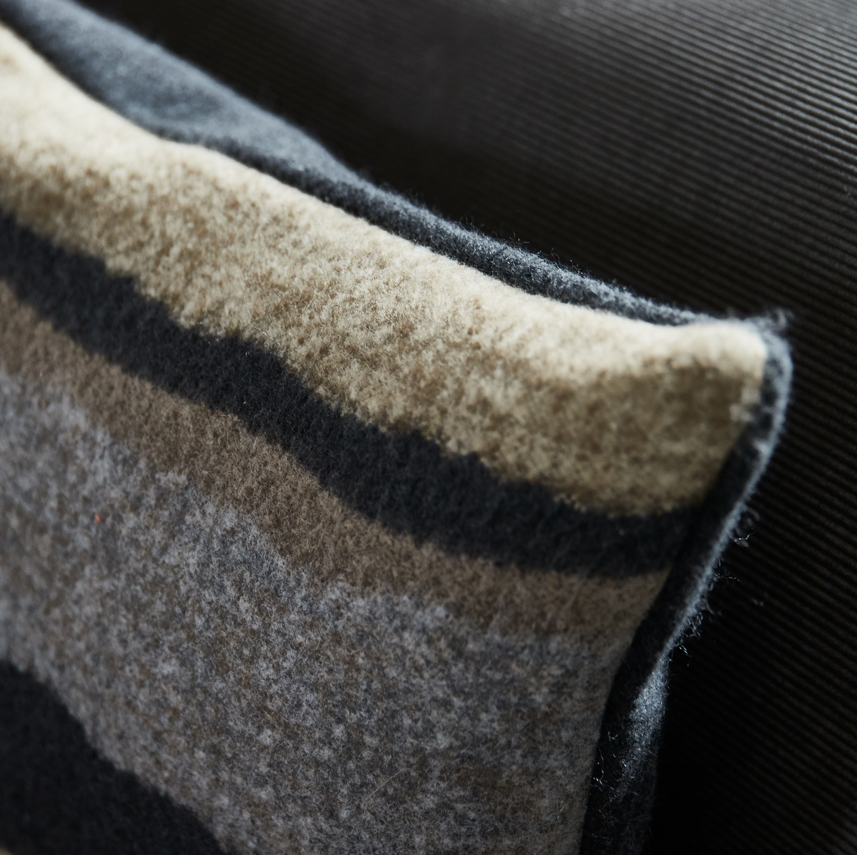 Nærbillede af blød uldpude med grå, beige og sorte striber mod mørk sofa.