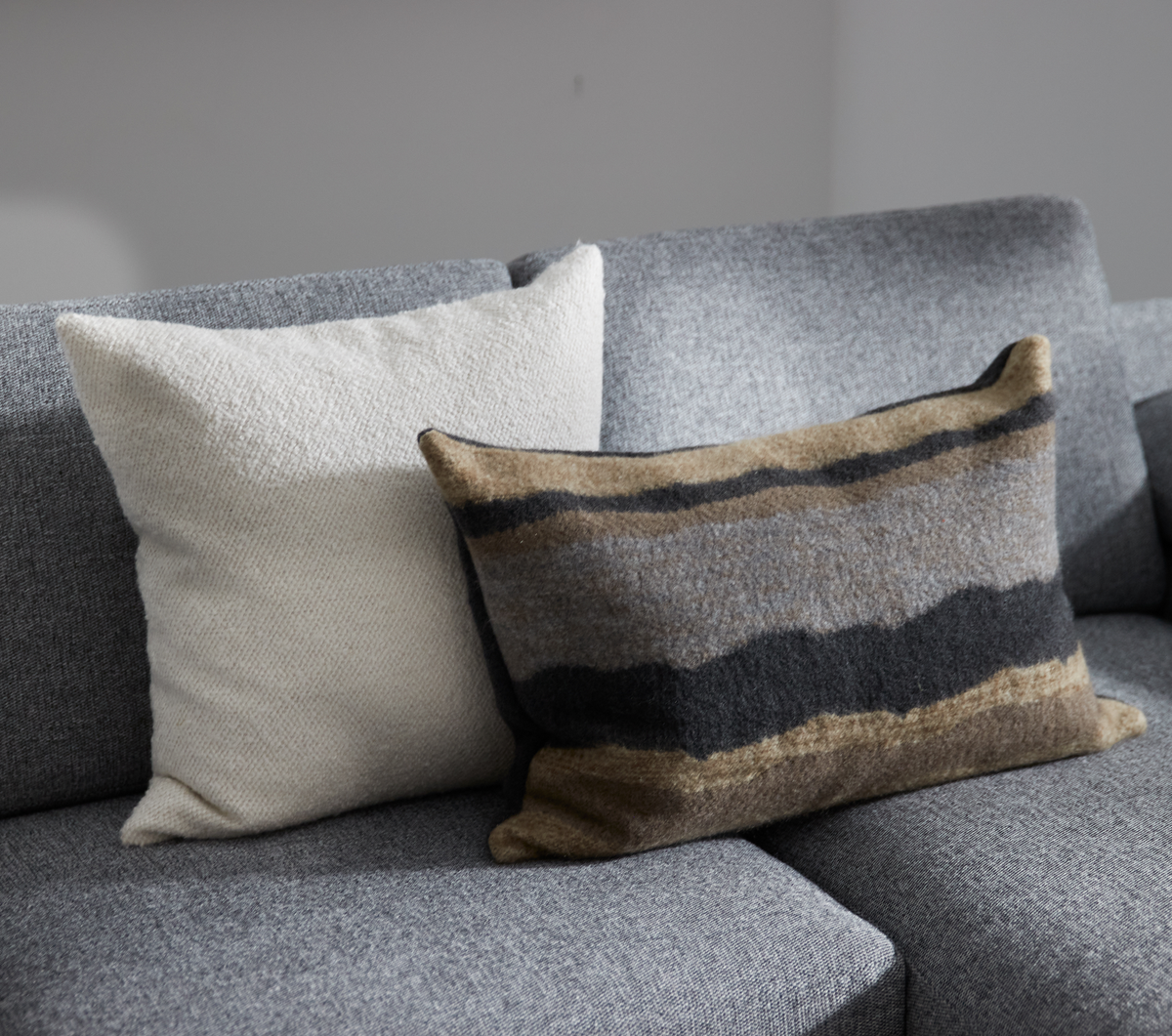 To pyntepuder på en grå sofa: en hvid firkantet og en rektangulær med grå‑brune jordfarvede striber.