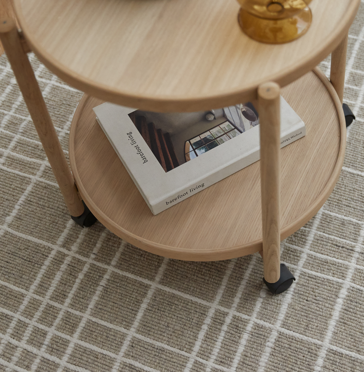 Nærbillede af et rundt sidebord i lyst træ på hjul med et ravfarvet glas og bogen ”barefoot living” på den nederste hylde på et beige ternet tæppe.