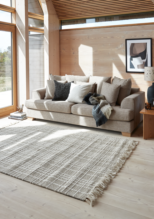 Lyst skandinavisk stue med beige sofa, neutrale puder og plaid, stor frynset tæppe på lyst trægulv, træbeklædte vægge, store vinduer, sidebord med keramiklampe og indramet abstrakt kunst.