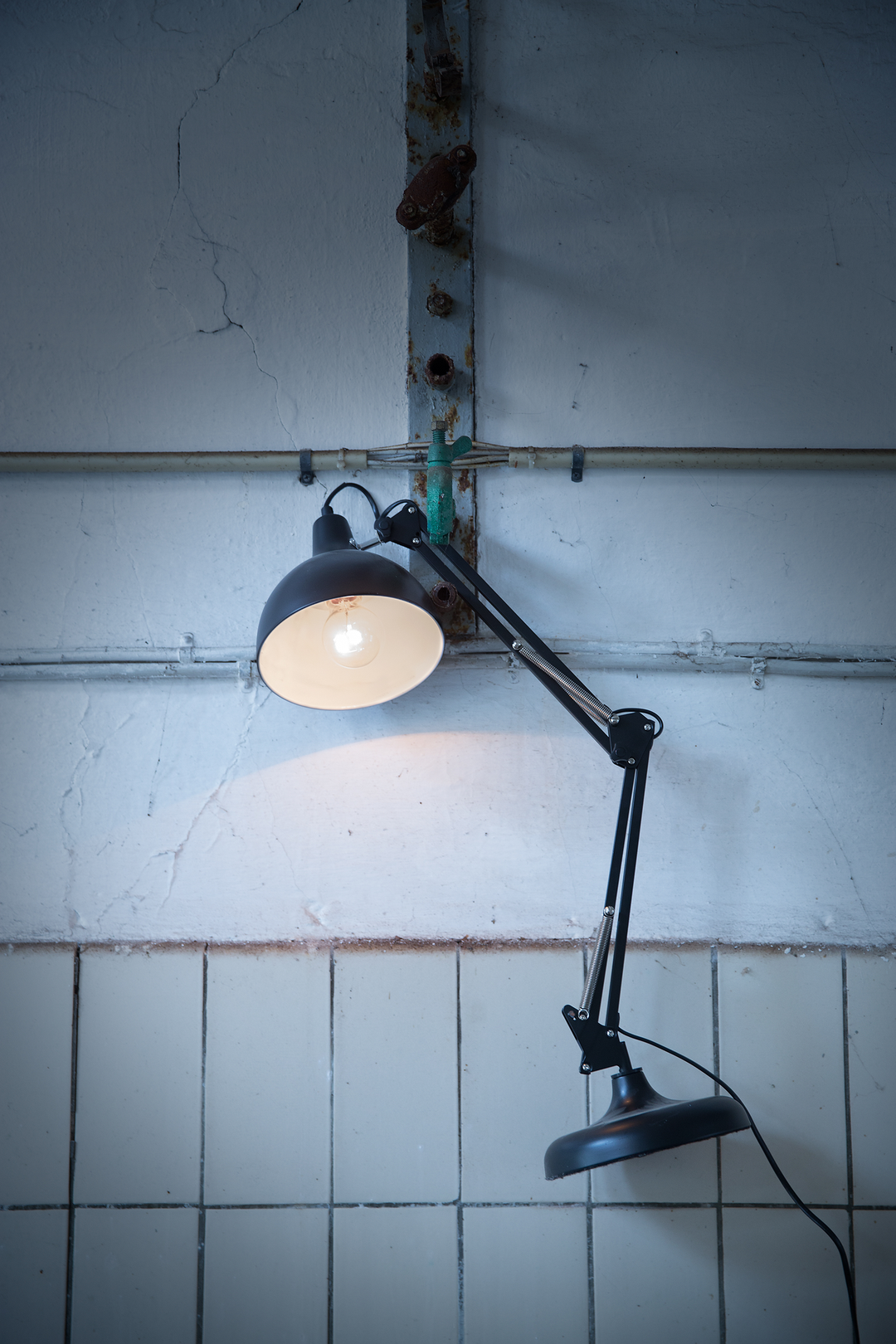 Industriel væg med rør og sort to-leddet arbejdslampe; den ene skærm er tændt og lyser mod den sprukne puds over fliser.