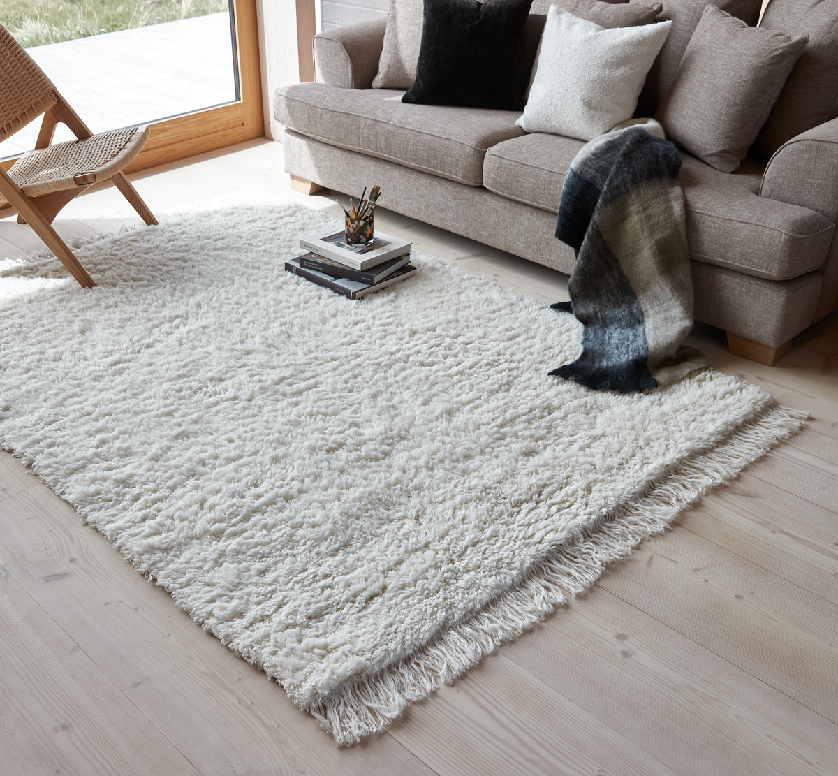 Lys stue med beige sofa, puder og plaid, stor hvid ryatæppe med frynser på lyst trægulv; bøger ligger på tæppet ved et stort vindue.