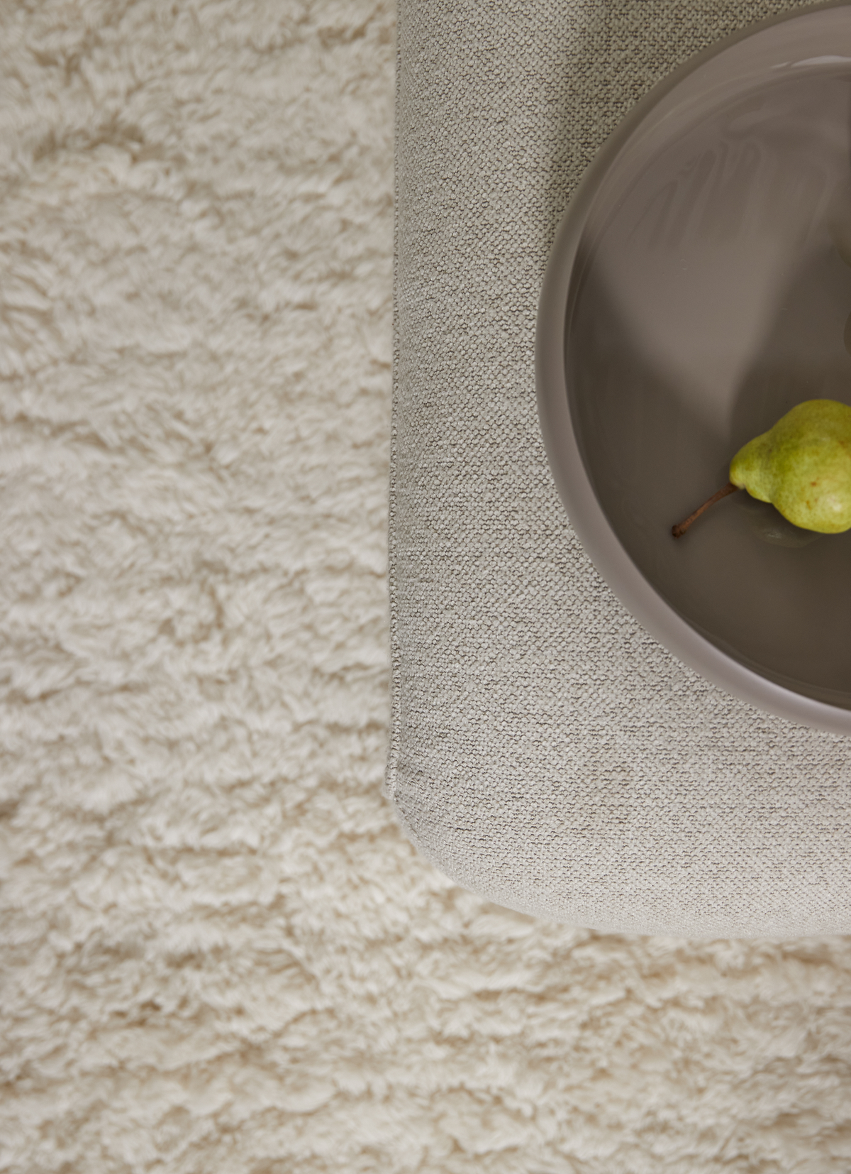 Nærbillede oppefra af beige sofa/puf med en taupefarvet tallerken med en grøn pære på et blødt cremefarvet tæppe.