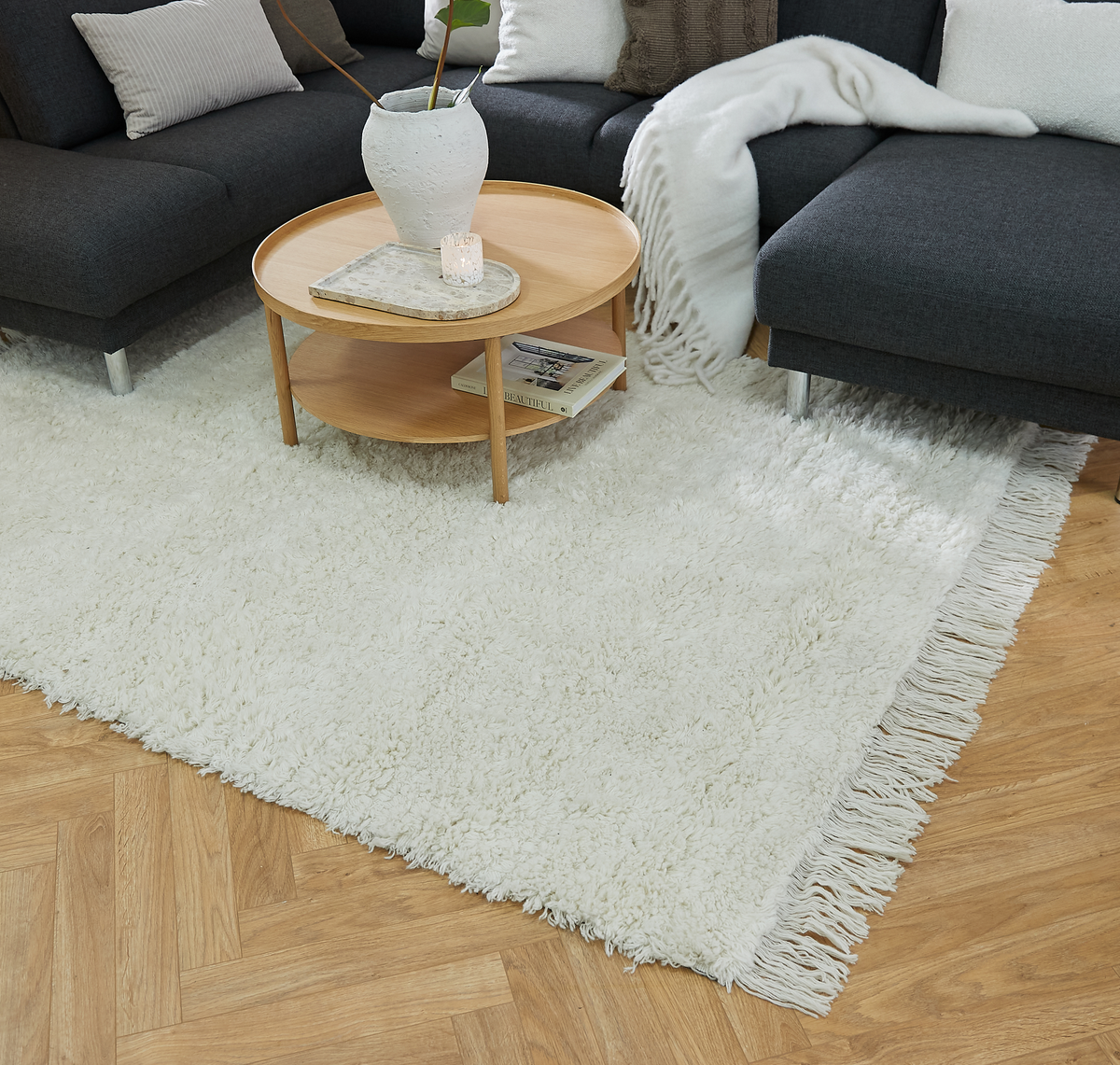 Lyst stue med hvid ryatæppe med frynser, rundt sofabord i træ med vase, lille lys og bogen “Beautiful”, ved en mørkegrå sofa med puder og plaid.