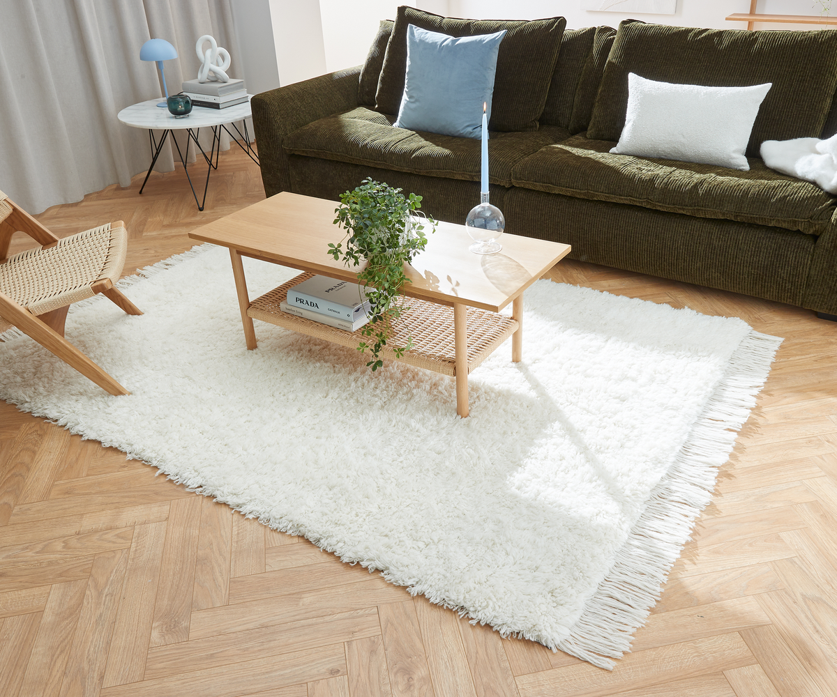 Lys stue med hvid, blød tæppe og lavt træbord med grøn plante, lyseblåt stearinlys i glasstage og bøger – en med titlen PRADA – foran en mørkegrøn sofa med blå og hvide puder.