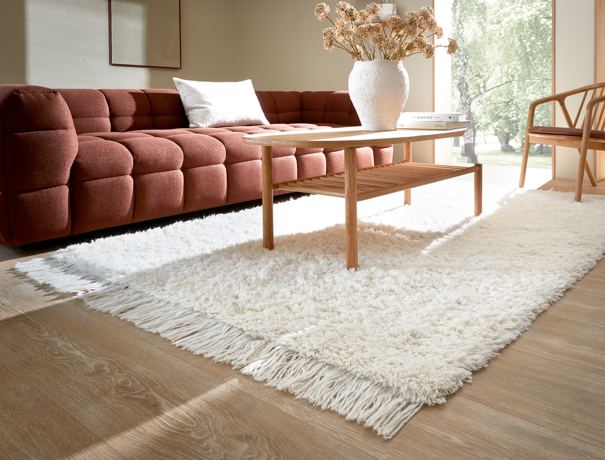 Lys stue med rustfarvet sofa, træsofabord med vase og bogen Kinfolk samt hvidt ryatæppe med frynser på lyst trægulv.