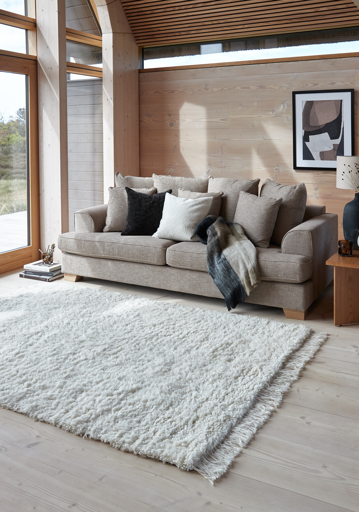 Lyst skandinavisk stue med beige sofa, puder og plaid, stort hvidt shaggy tæppe på lyst trægulv ved store vinduer, træbord med keramiklampe og abstrakt vægkunst.