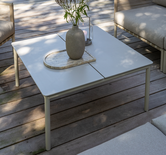 Lavt kvadratisk udendørsbord i lysegrå metal på en træterrasse med en vase med grønne grene, en stenbakke og en glaslanterne med tændt lys, omgivet af lyse loungestole.