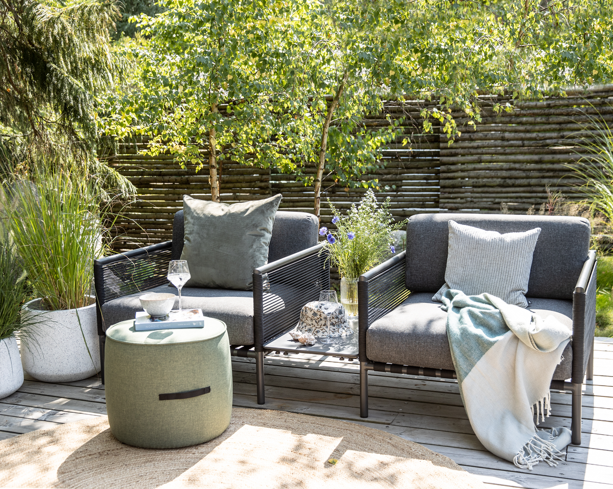 To grå udestole med puder og plaid på en træterrasse; en grøn puff bruges som bord med glas og skål, omgivet af krukker med planter og et træhegn i en solrig have.