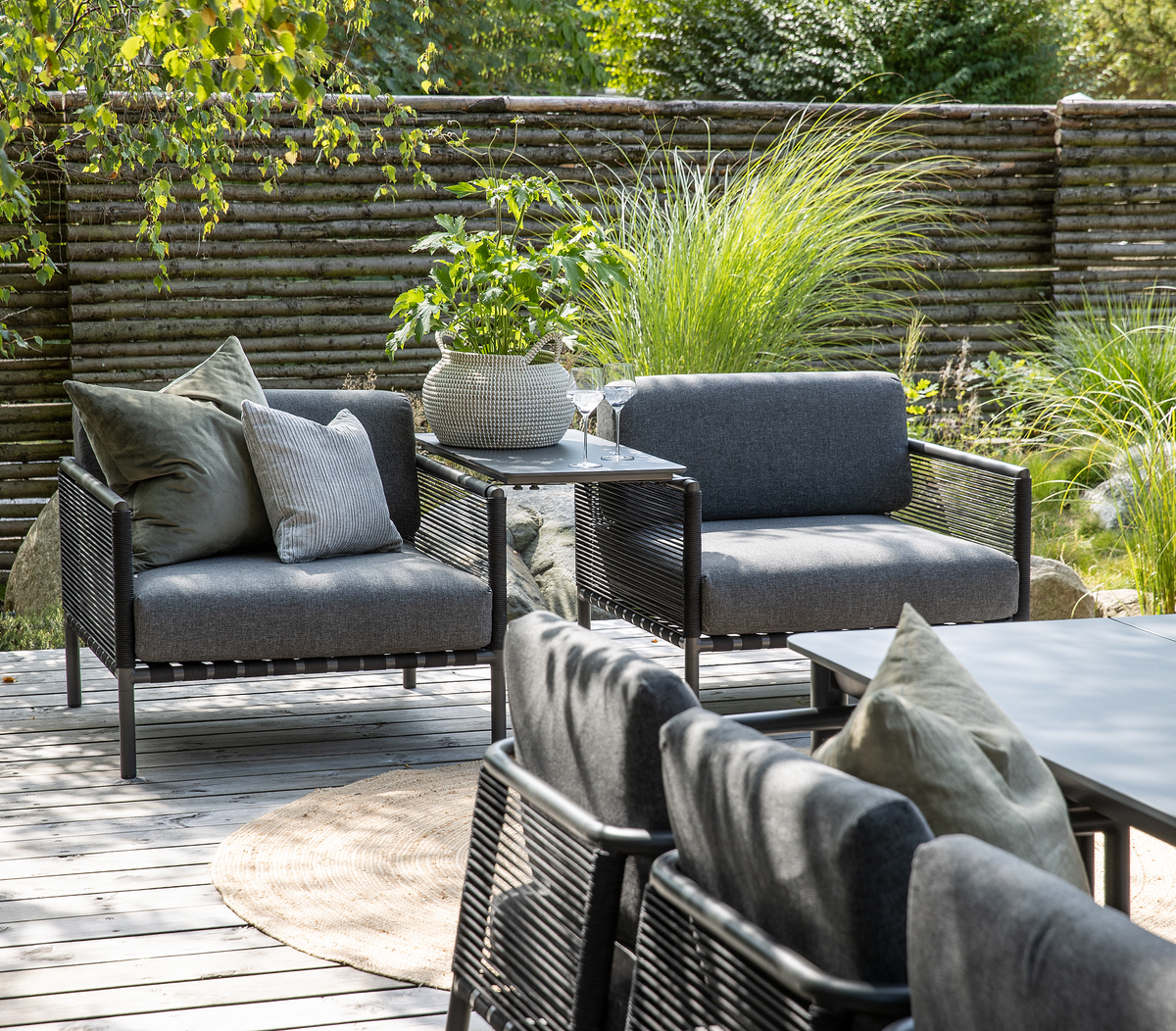 Moderne udestue-lounge med grå reb-lænestole, lille bord med potteplante og vinglas på en træterrasse i haven, omgivet af prydgræs og træhegn i dagslys.