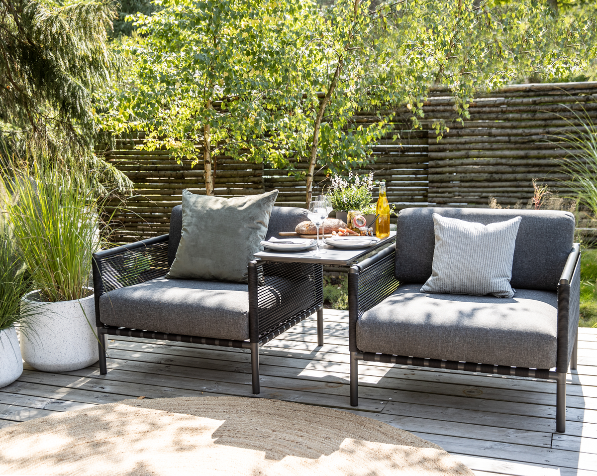 Terrasse med to mørkegrå loungestole og et lille bord på trædæk, med puder, potteplanter samt brød og drikke i sollys foran et træhegn.