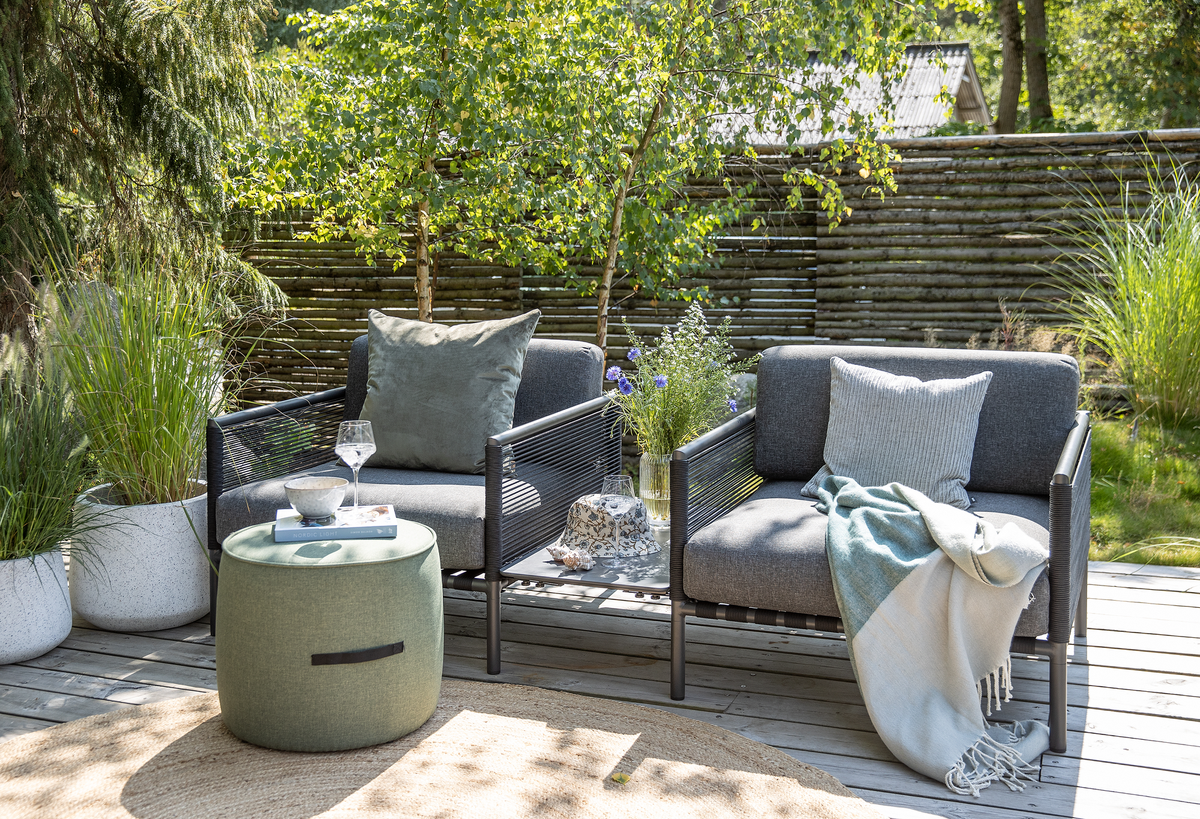 To grå udestole på en træterrasse med en grøn puf som bord med vinglas og skål, puder og plaid på stolene samt en vase med sommerblomster; krukkeplanter og træhegn i en solrig have.