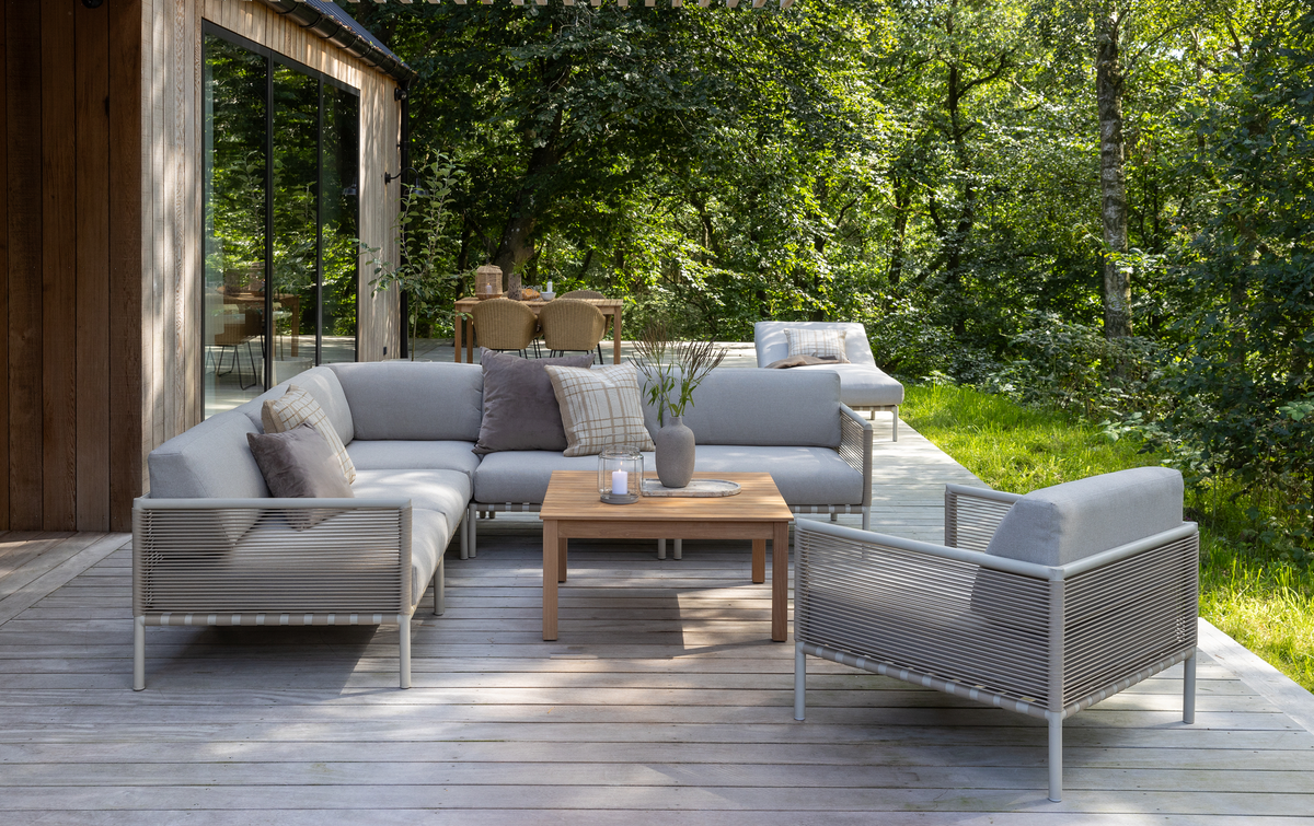 Lysgrå modulsofa med matchende lænestol og træ-sofabord på en terrasse; vase med grene og et lys på bordet, skov i baggrunden i dagslys.