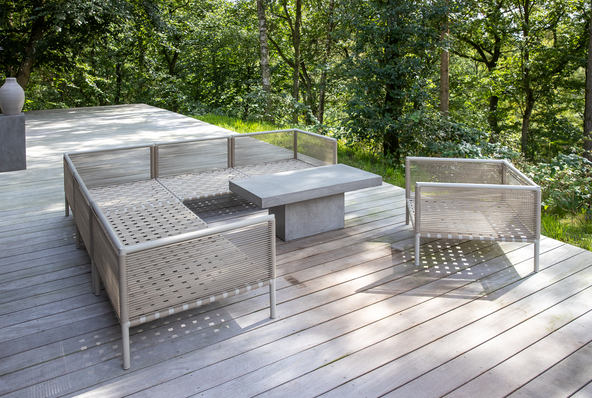 Udendørs terrasse med moderne loungemøbler i snor og et betonbord på trædæk, omgivet af grøn skov.