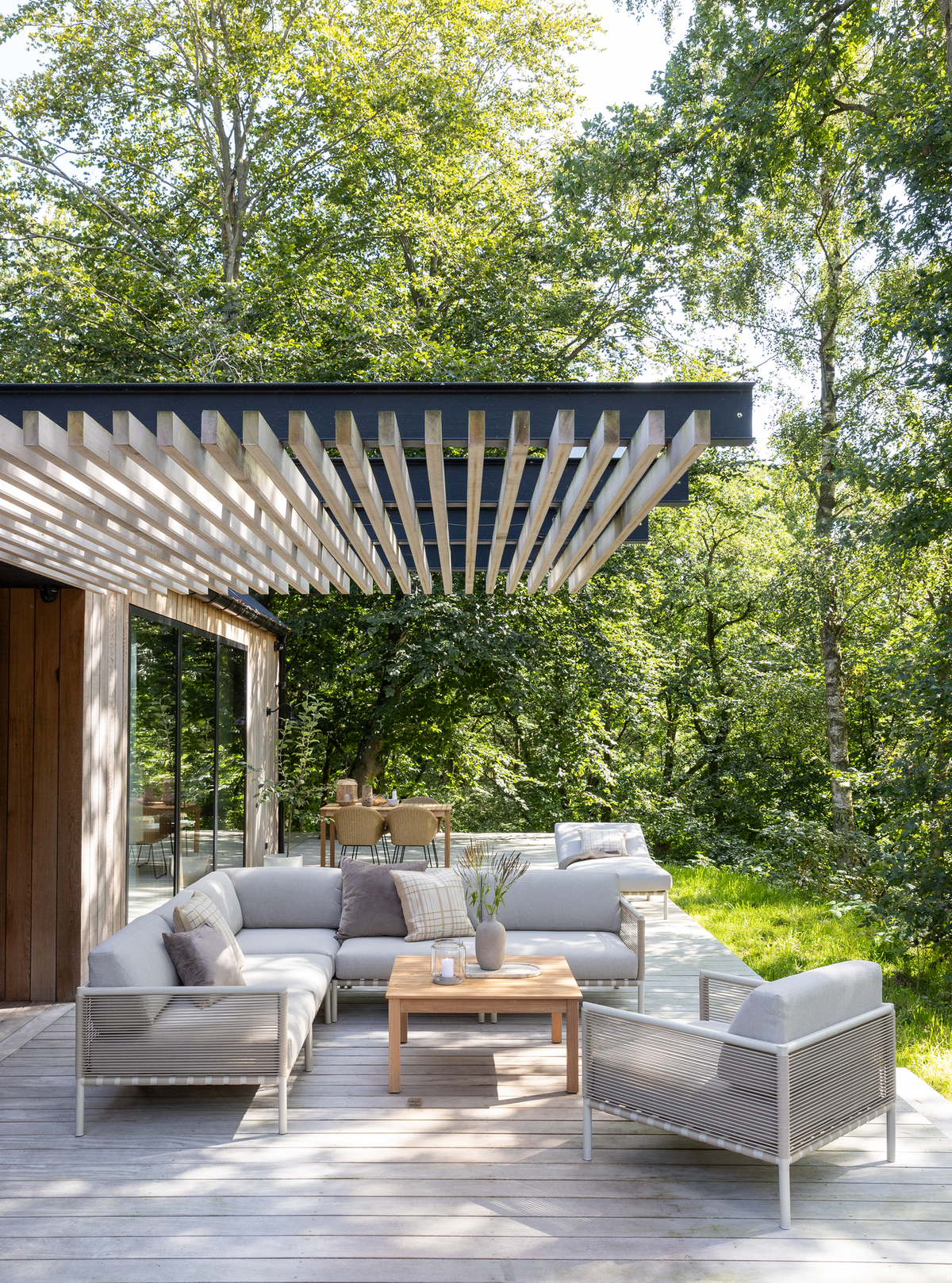 Lys terrasse med moderne grå loungesofa, lænestol og træbord under en pergola, omgivet af grøn skov i dagslys.