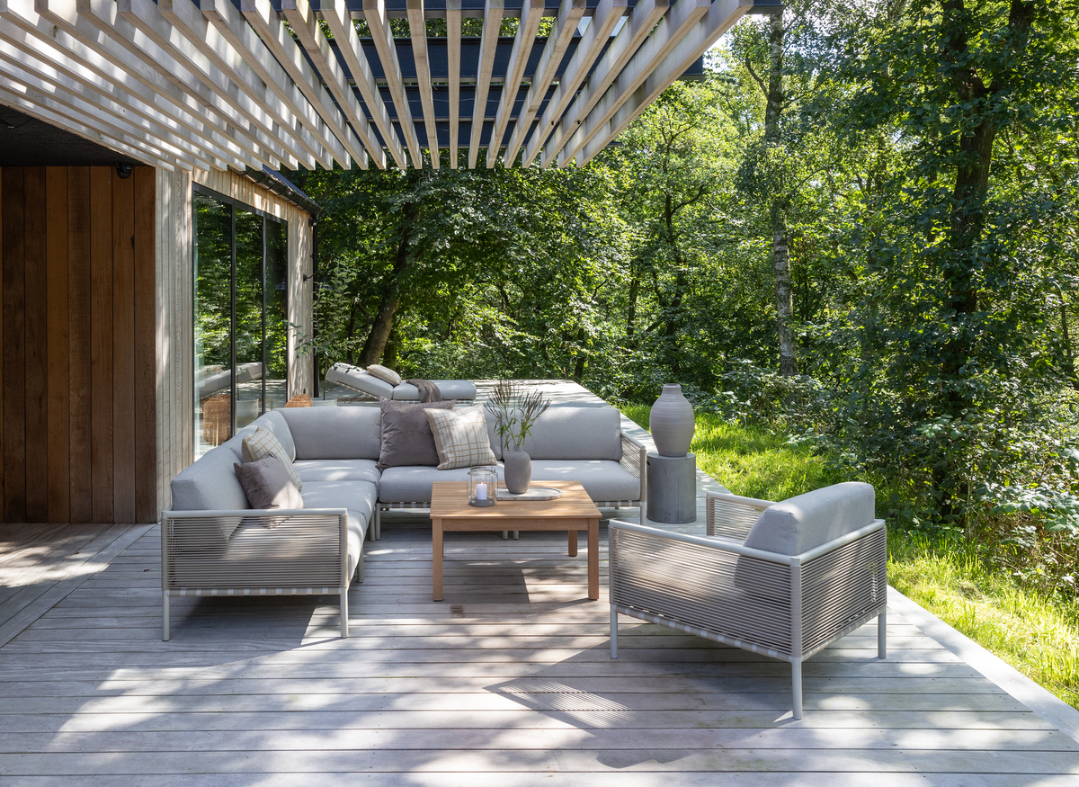 Lyst træterrasse med grå udemøbler under pergola, loungehjørne med puder og sofabord med vase og lys, omgivet af grøn skov.