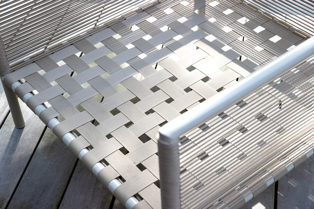 Nærbillede af en udendørs stol med lysegrå vævede bånd og aluminiumsramme på en træterrasse.