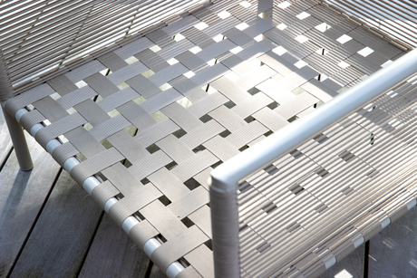 Nærbillede af en udendørs stol med lysegrå vævede bånd og aluminiumsramme på en træterrasse.