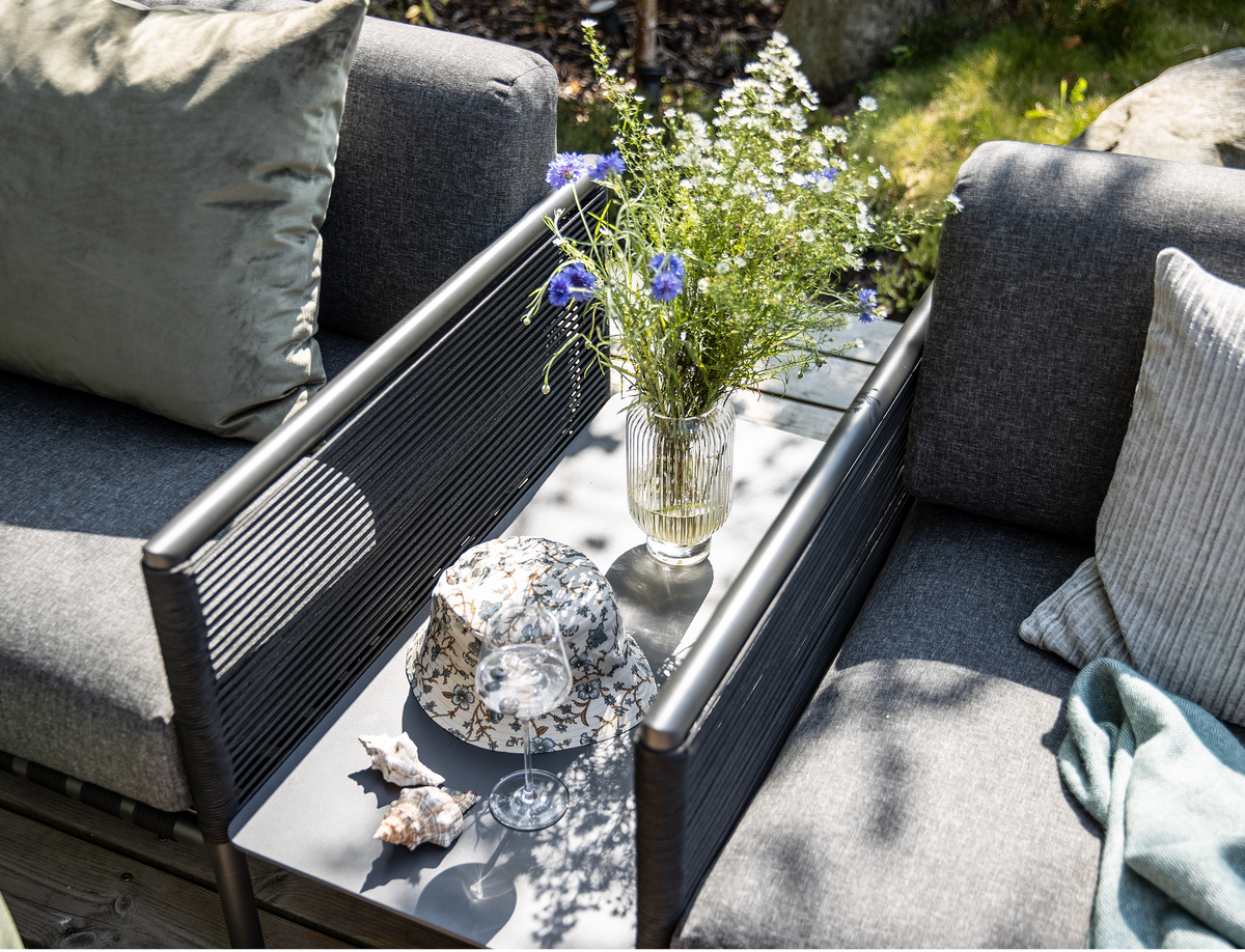 Udendørs sofasæt med grå hynder og et smalt bord med glasvase med sommerblomster, solhat, vinglas og strandskaller i sollys.