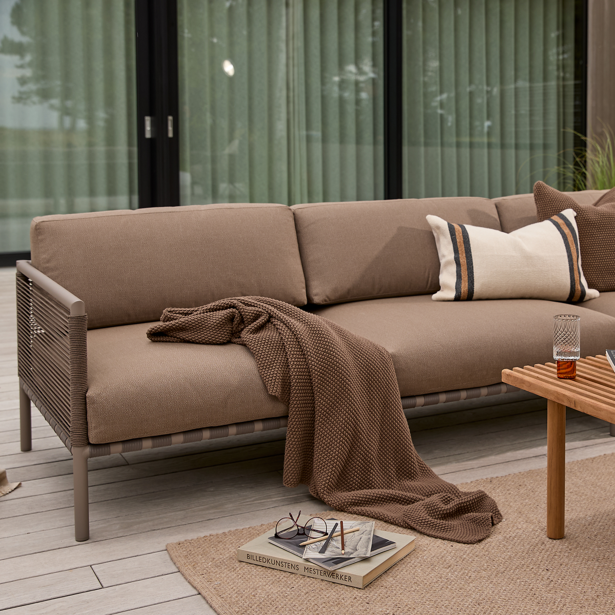 Brun udendørs sofa med plaid og puder på terrasse; glas på rillet træbord; bog BILLEDKUNSTENS MESTERVÆRKER på tæppet.