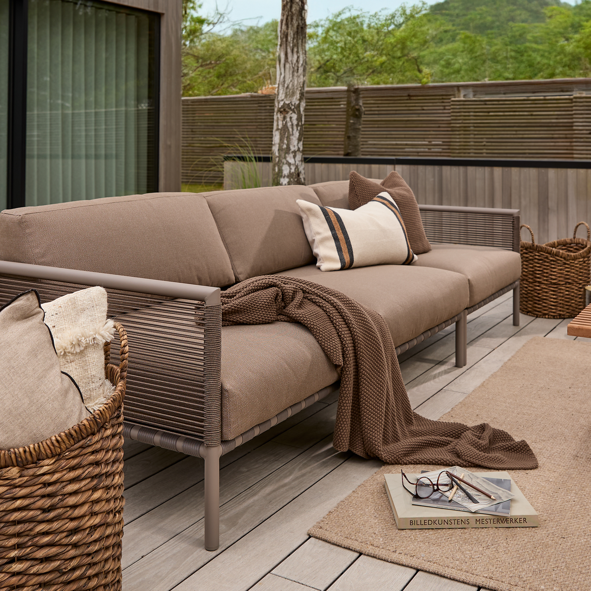 Udendørs sofa i beige med plaid og puder på terrasse; bakke med briller og bogen ”Billedkunstens Mesterværker” på tæppet.