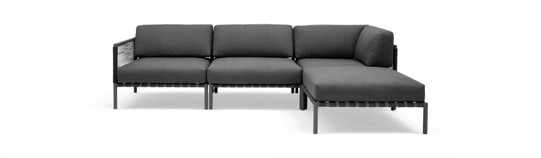 Mørkegrå modulsofa i L‑form med chaiselong til højre, fritstående mod sort baggrund.