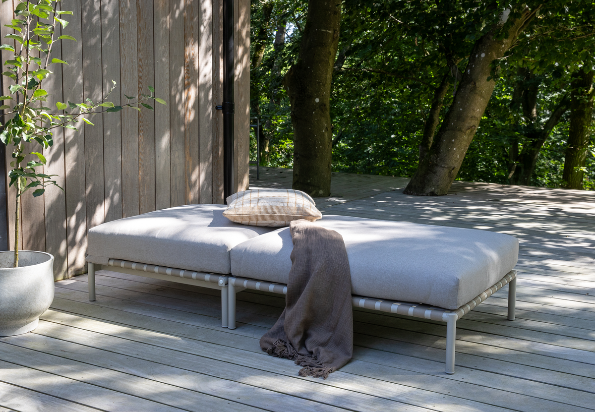Lysegrå udendørs daybed på træterrasse med pude og plaid ved træfacade i træernes skygge.