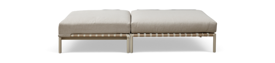 Modulær ottoman/bænk med to beige hynder på lys metalramme med vævet rembund, set forfra på mørk baggrund.