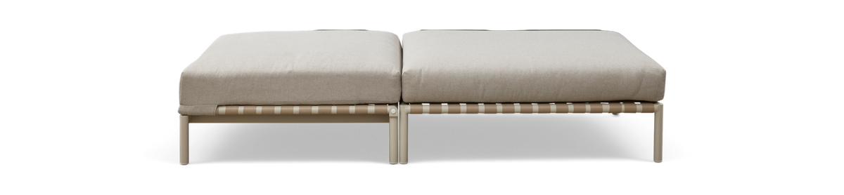 Modulær ottoman/bænk med to beige hynder på lys metalramme med vævet rembund, set forfra på mørk baggrund.