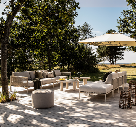 Lys træterrasse med beige loungesofaer, runde sofaborde og stor parasol omgivet af grønt.