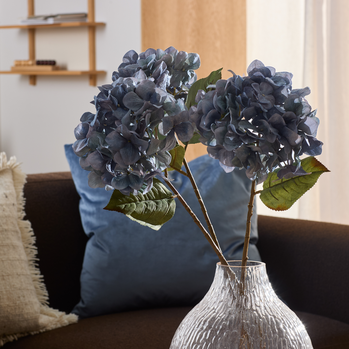 Blå hortensia i klar, struktureret glasvase på sofa med blå pude i en lys stue.