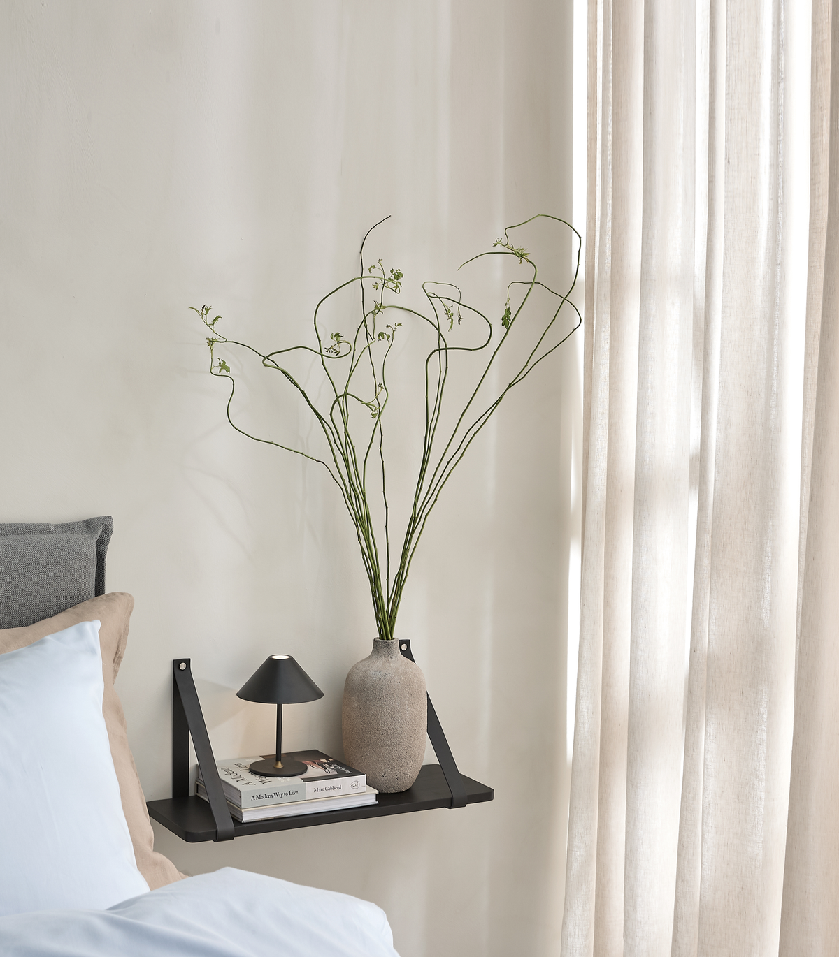 Lyst soveværelse med sort væghylde som sengebord, lille sort bordlampe, bøger og beige vase med høje grønne grene; linnedgardiner i dagslys.