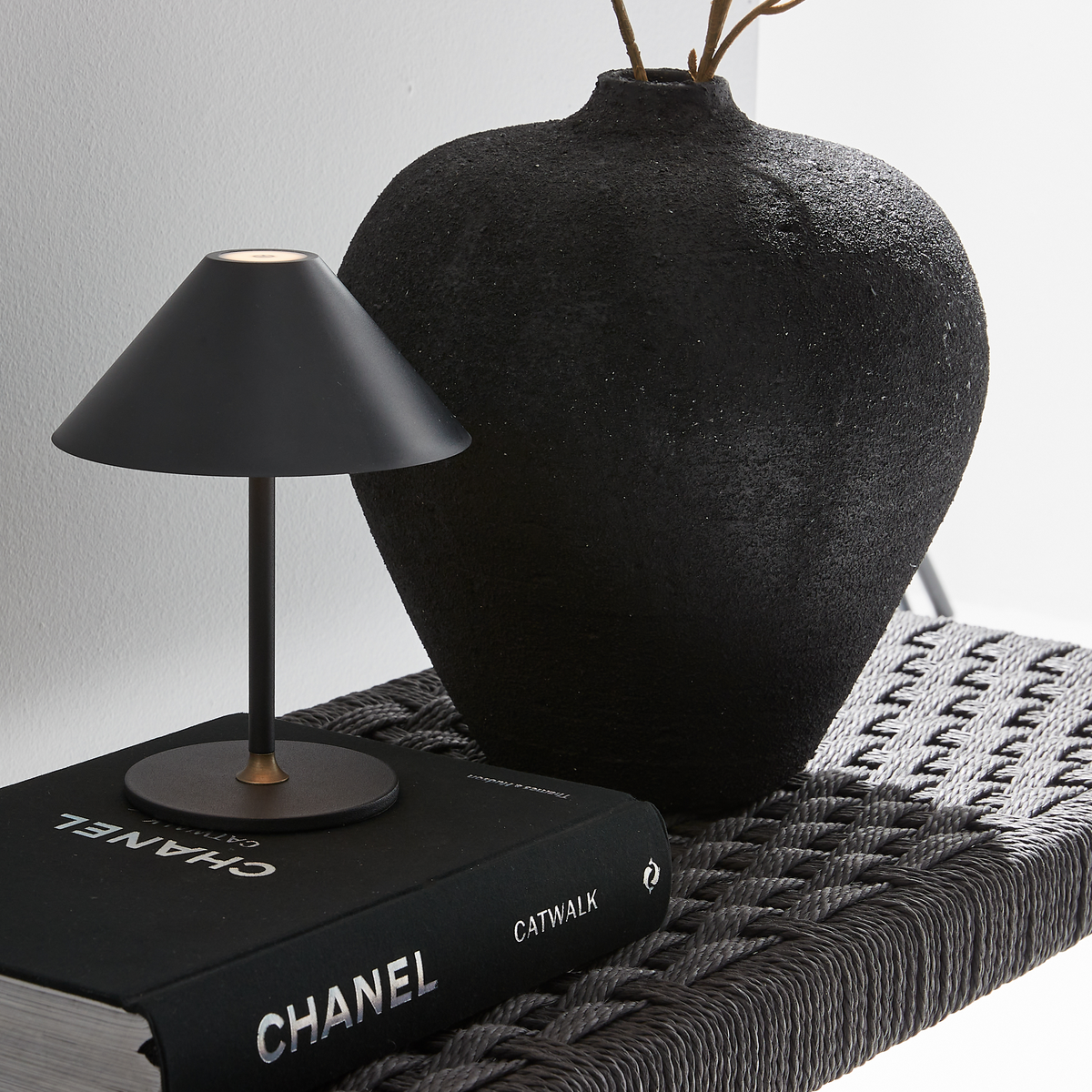 Lille sort bordlampe står på bogen Chanel Catwalk ved siden af en stor mat sort vase på en fletreol i et lyst rum.
